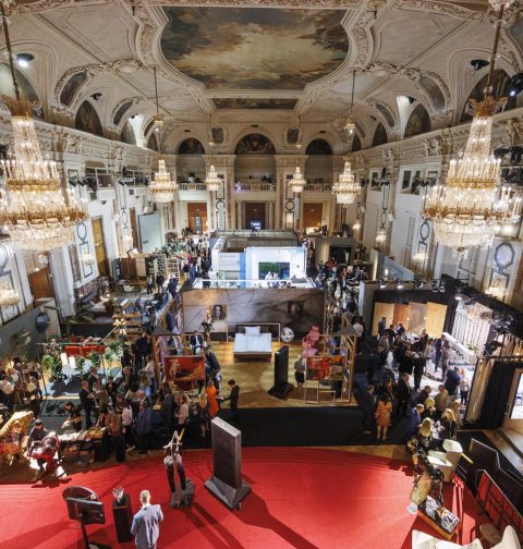 prunkvoller Raum der Hofburg mit Messeständen und Besuchern, Kronleuchter, roter Boden, Gemälde an der Decke