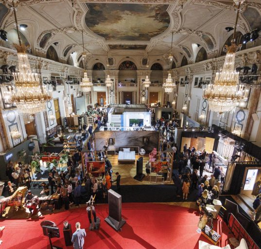 prunkvoller Raum der Hofburg mit Messeständen und Besuchern, Kronleuchter, roter Boden, Gemälde an der Decke