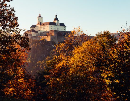 Burg Forrchtenstein thront am Hügel vor orange-braunen Baumkronen. Der Himmel ist hellblau. herbstliche Stimmung