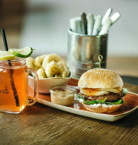 Burger von Le Berger mit Limonade auf Holztisch. Im Hintergrund Besteck in einem Metallgefäss.