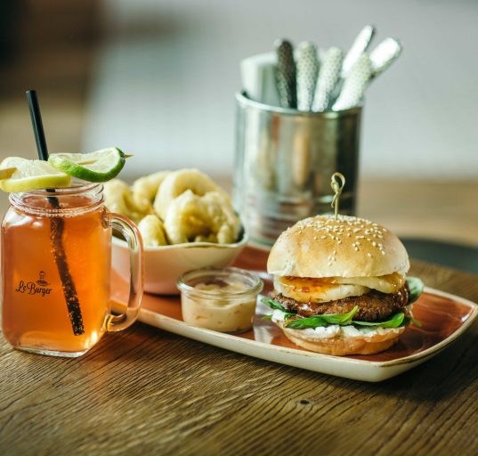 Burger von Le Berger mit Limonade auf Holztisch. Im Hintergrund Besteck in einem Metallgefäss.