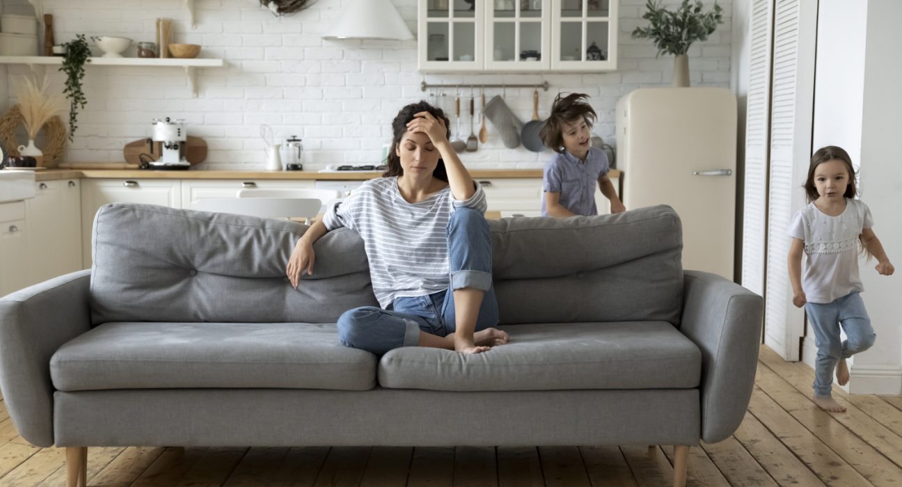 Frau sitzt gestresst auf Couch, um sie herum laufen zwei Kinder