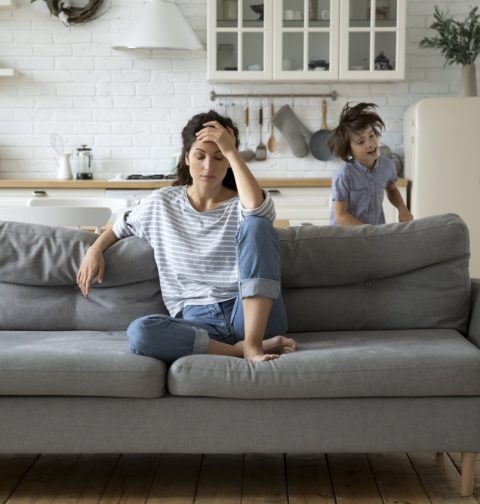 Frau sitzt gestresst auf Couch, um sie herum laufen zwei Kinder
