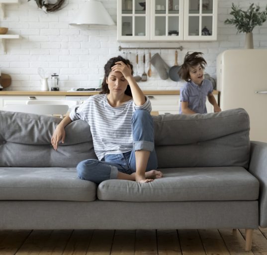 Frau sitzt gestresst auf Couch, um sie herum laufen zwei Kinder