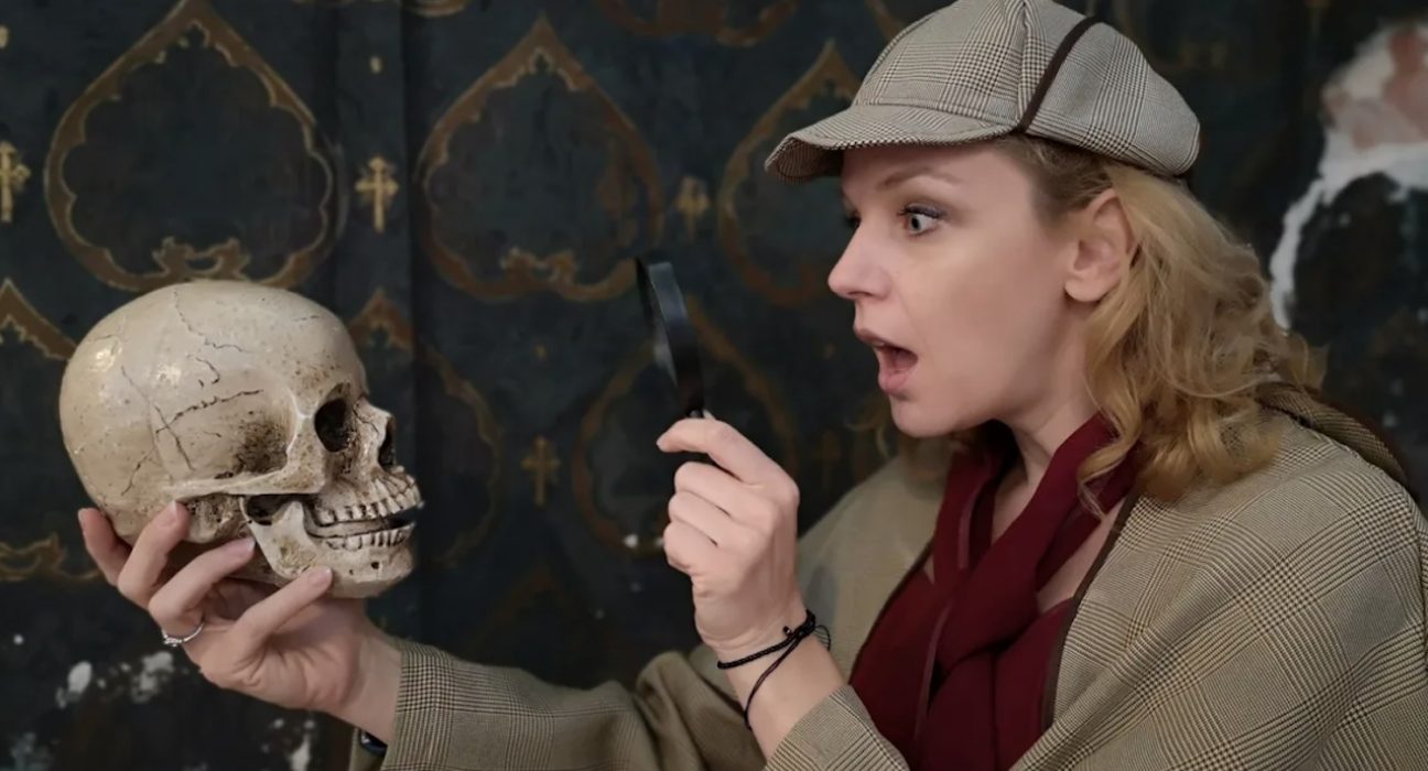 Eine junge Frau mit Sherlock-Homes-Mütze und einem Vergrößerungsglas untersucht einen Totenkopf. (Gruselführung!) @ esterhazy.at