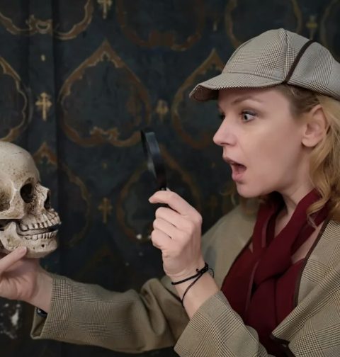 Eine junge Frau mit Sherlock-Homes-Mütze und einem Vergrößerungsglas untersucht einen Totenkopf. (Gruselführung!) @ esterhazy.at