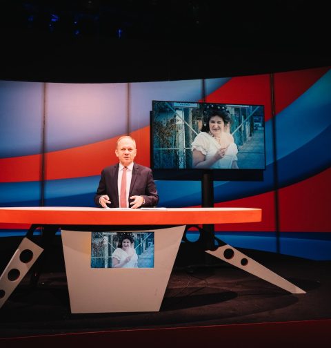 Der Moderator Paul Kraker in einem TV-Studio (der Tagespresse), auf einer Leinwand im Hintergrund ist die Schauspielerin Maria Hofstätter in einem weißen Kleid zu sehen. @ Rabenhof/Ingo Pertramer