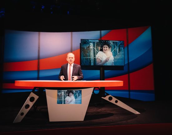 Der Moderator Paul Kraker in einem TV-Studio (der Tagespresse), auf einer Leinwand im Hintergrund ist die Schauspielerin Maria Hofstätter in einem weißen Kleid zu sehen. @ Rabenhof/Ingo Pertramer