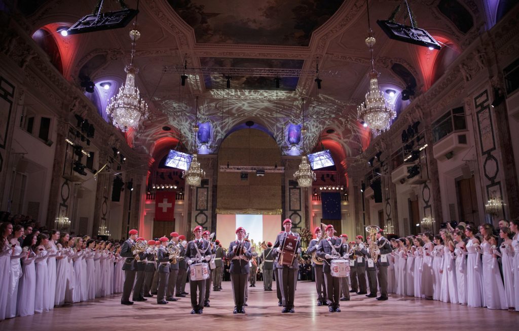 Festsaal mit Kronleuchtern, an den Bildrändern stehen Damen in weißem Ballkleid, in der Mitte spielt eine Militärkapelle