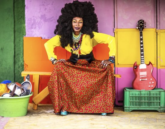 Fatoumata Diawara steht vor einer bunten Wand, links von ihr ein Plastiktrog mit Geschirr, rechts eine Gitarre, die auf einer Kiste steht. © Alun Be