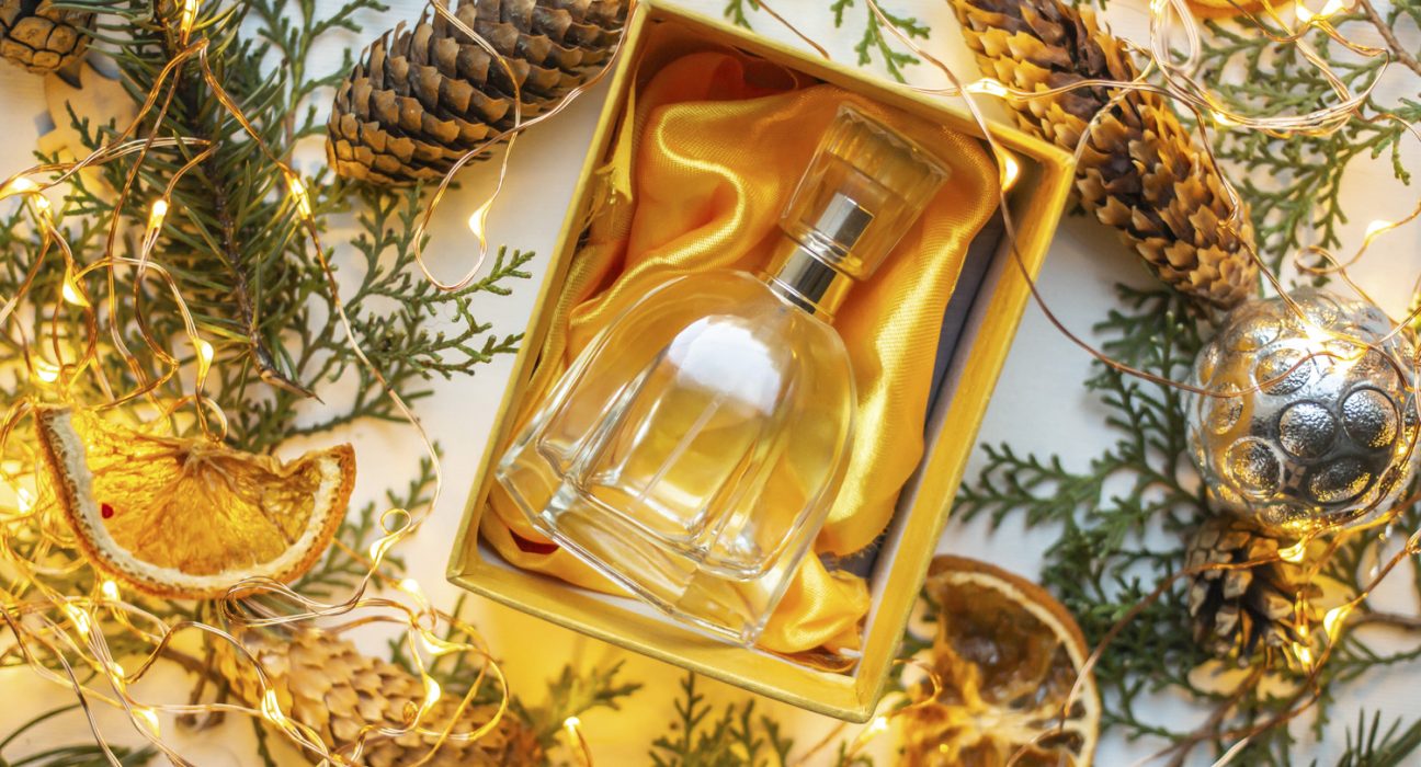 Parfumflasche in Box auf orangem Tuch, ringsum Dekoration aus Tannenzapfen, Blättern, getrockneten Orangen und Lichterkette