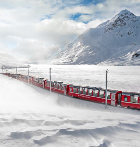 Roter Zug Bernina Express fährt durch ein schneebedecktes Tal beim Lago Bianco in der Schweiz
