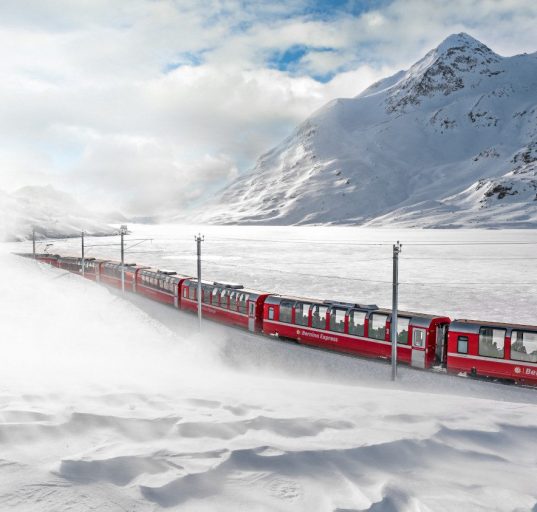 Roter Zug Bernina Express fährt durch ein schneebedecktes Tal beim Lago Bianco in der Schweiz