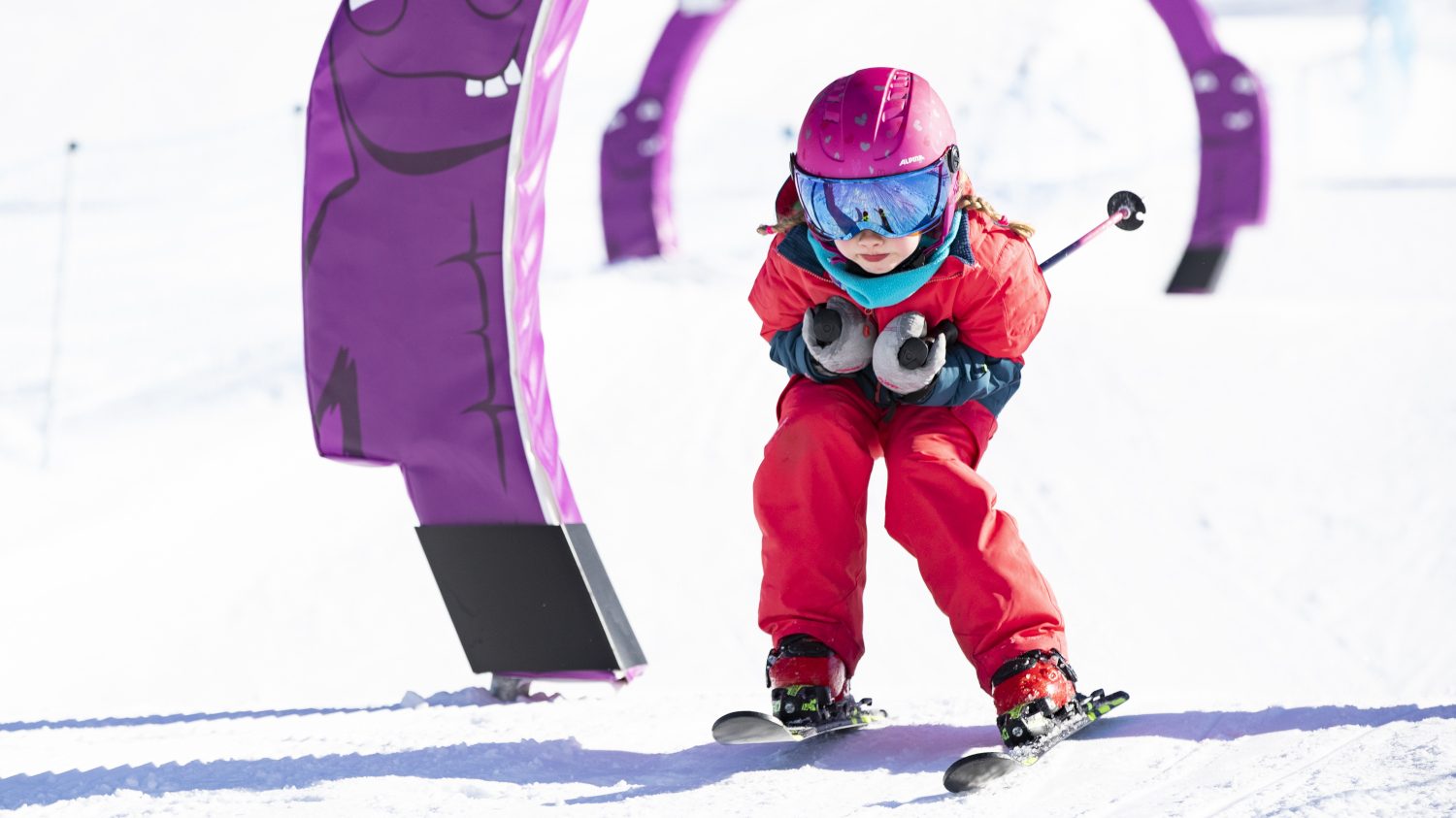 Kleinkind fährt auf Skiern im Schuss durch lila Torbögen im Familypark des Skicircus Saalbach-Hinterglemm