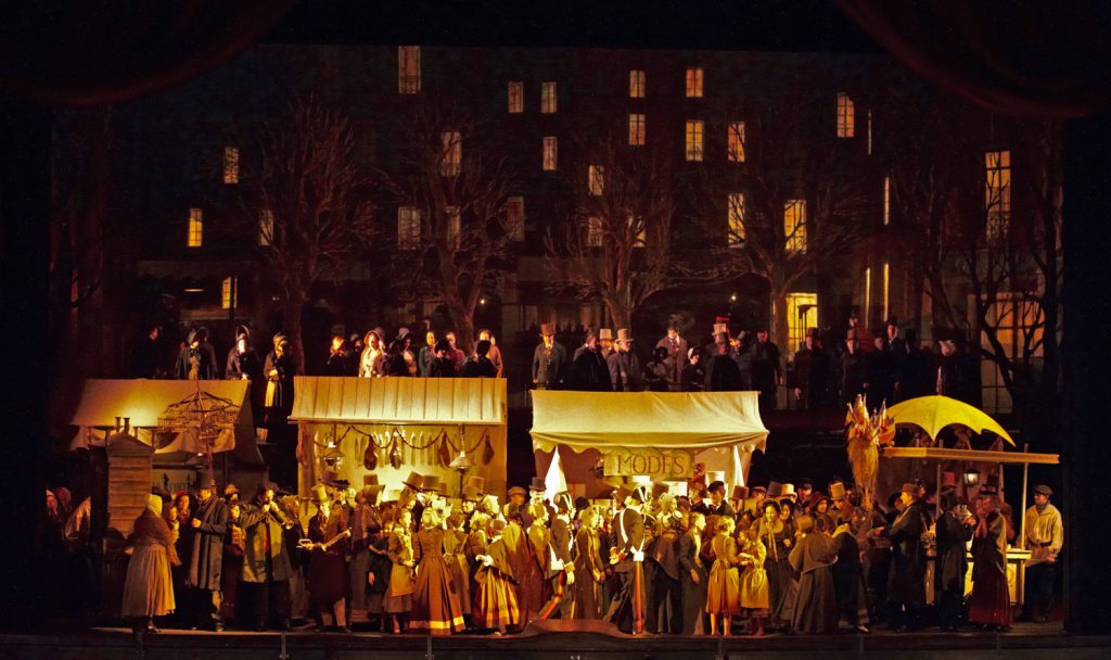 Bühnenbild von La Boheme. Im Hintergrund Häuser mit beleuchteten Fenstern. Viele Menschen, Marktstände im Vordergrund