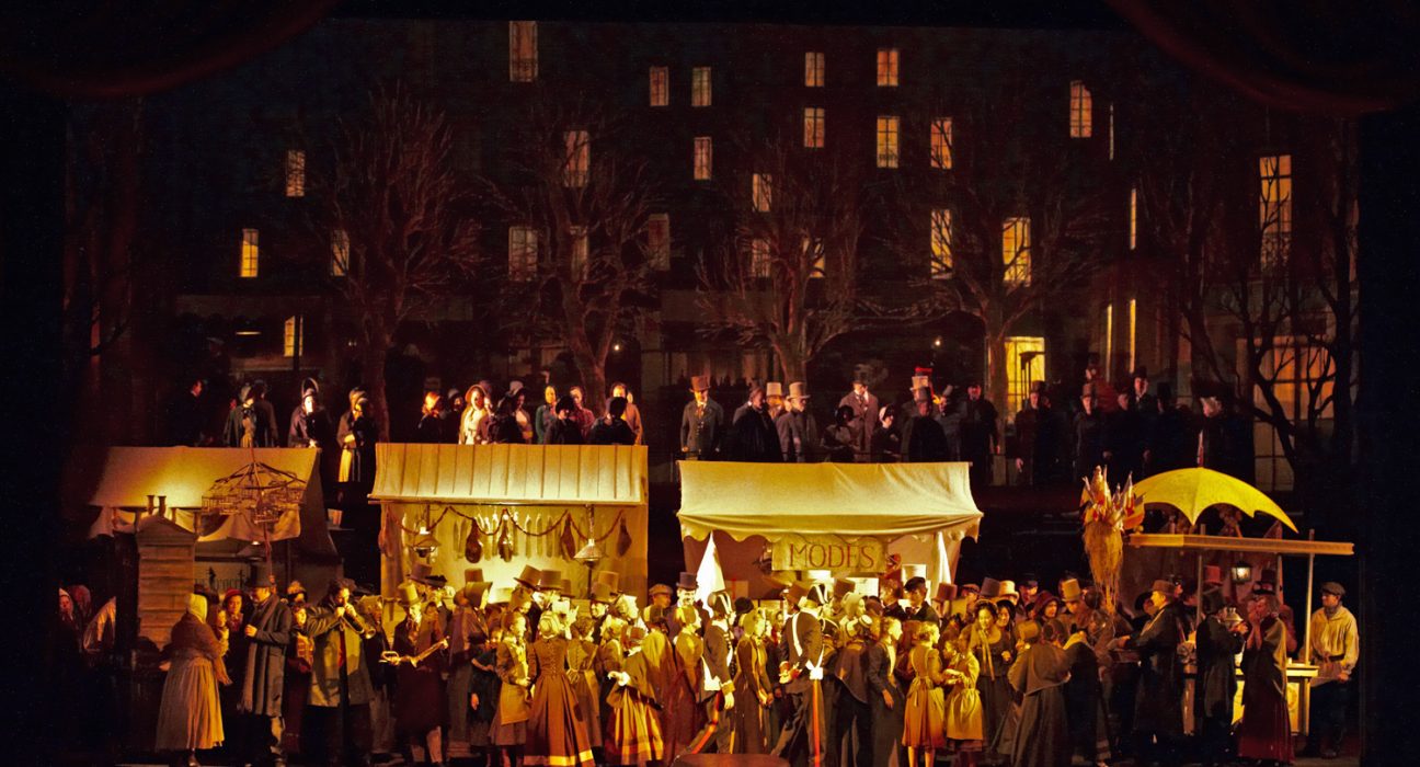 Bühnenbild von La Boheme. Im Hintergrund Häuser mit beleuchteten Fenstern. Viele Menschen, Marktstände im Vordergrund