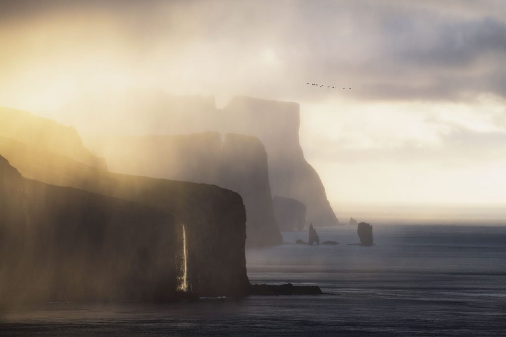 Blick vom Meer aus auf den Sonnenuntergang hinter verregneter Küste der Färöer-Inseln