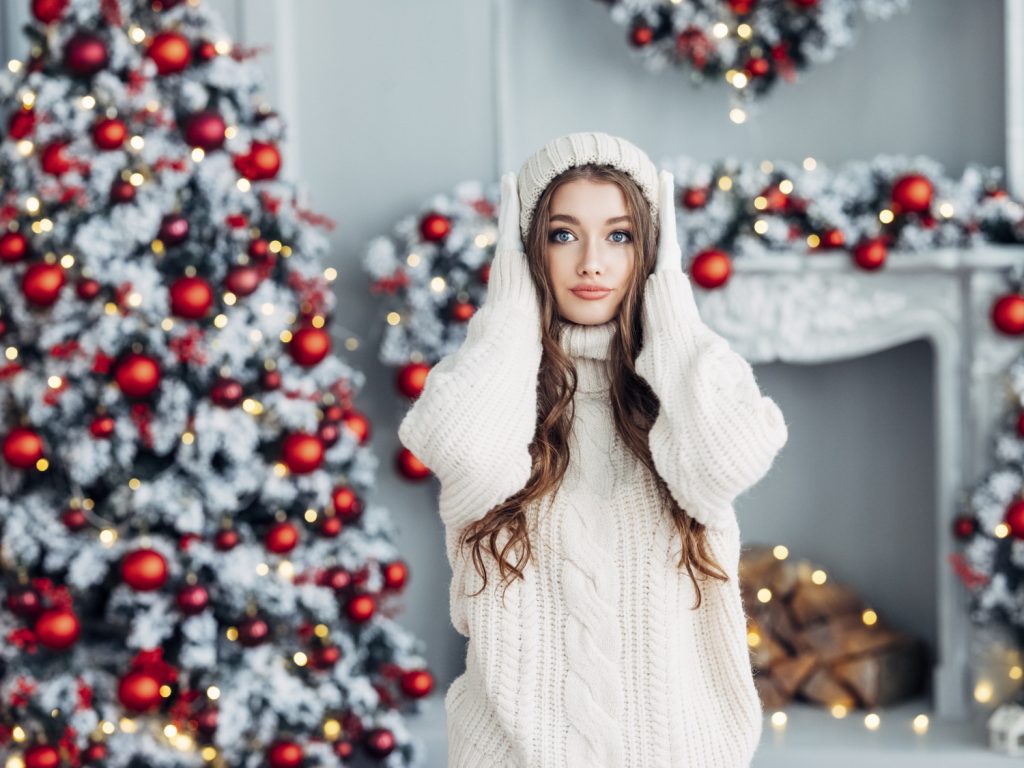 Weihnachtsstress: Frau mit Pullover, Handschuhen und Haube in creme vor weißen Weihnachtsbaum mit roten Kugeln.