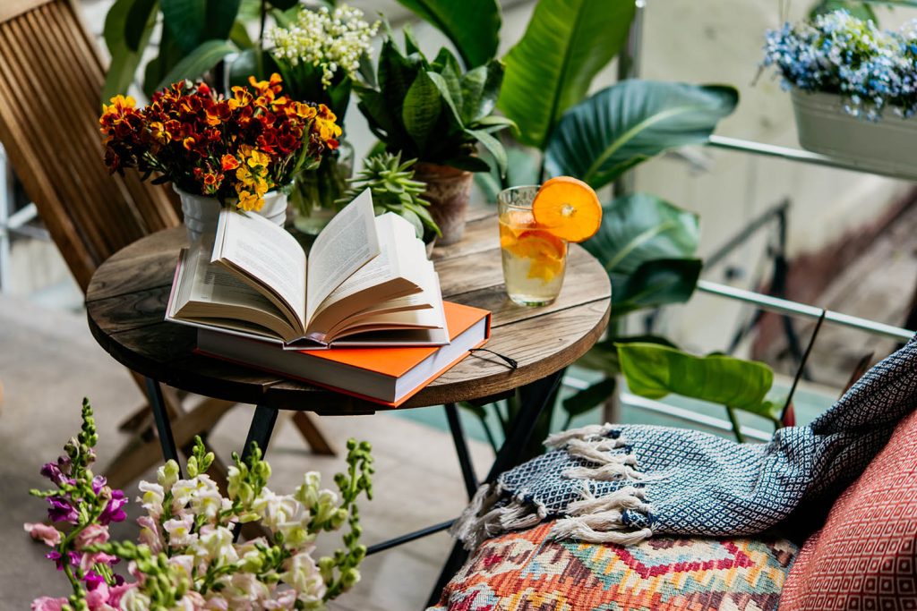 Trends im Garten im Frühling: Aufgeschlagenes Buch auf Holztisch mit Getränk in Glas mit Orangenscheiben und Blumen-Deko rundherum.