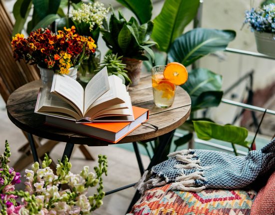 Trends im Garten im Frühling: Aufgeschlagenes Buch auf Holztisch mit Getränk in Glas mit Orangenscheiben und Blumen-Deko rundherum.