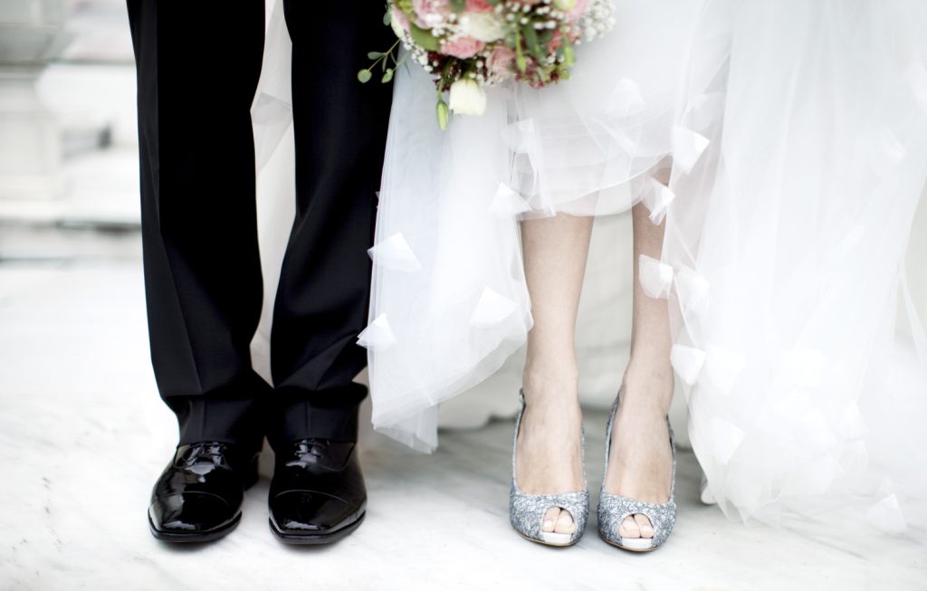 Hochzeitsstil: Braut und Bräutigam nebeneinander. Nur Schuhe, Hose und Kleid sind zu sehen. Ein Blumenstrauß ist am oberen Ende zu sehen.