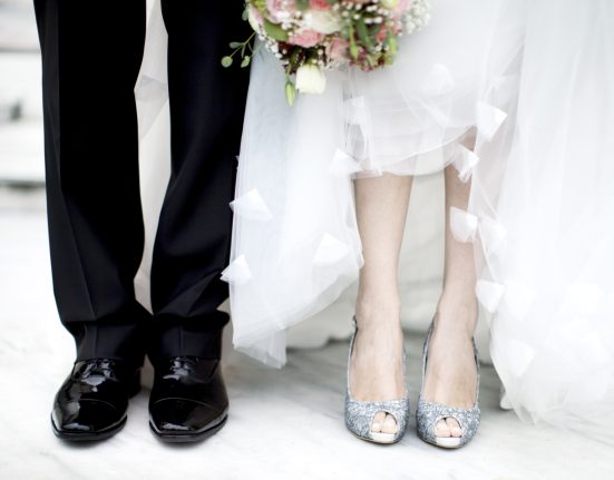 Hochzeitsstil: Braut und Bräutigam nebeneinander. Nur Schuhe, Hose und Kleid sind zu sehen. Ein Blumenstrauß ist am oberen Ende zu sehen.