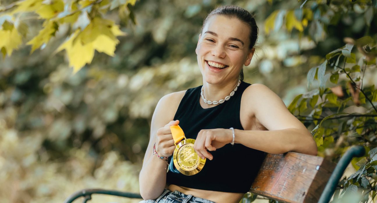 Eine junge Frau sitzt auf einer Parkbank und hält eine Goldmedaille in der Hand.