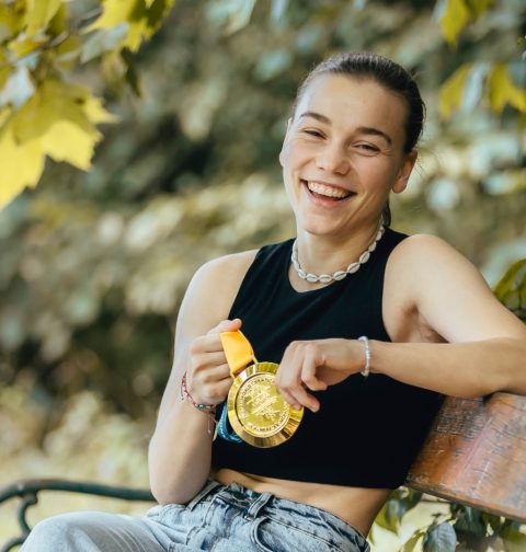 Eine junge Frau sitzt auf einer Parkbank und hält eine Goldmedaille in der Hand.