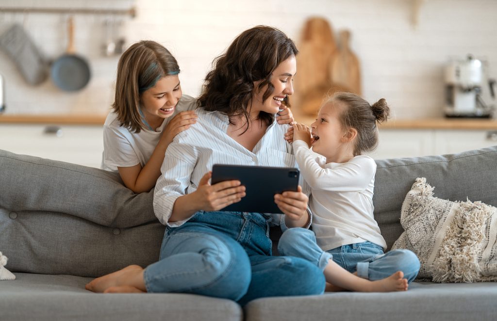 Medienkompetenz bei Kindern fördern: Mutter mit Tablet in der Hand und zwei Töchtern auf der Couch.