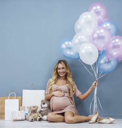 Babyparty: Schwangere Frau mit Geschenken und Luftballons in der Hand vor blauer Wand.