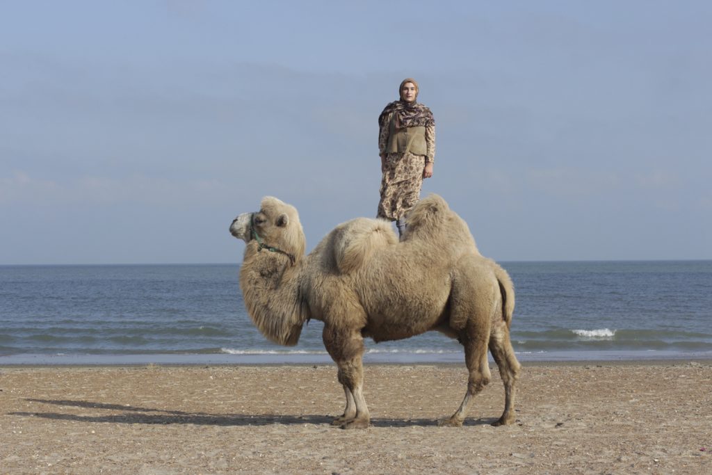 Frau steht am Strand auf einem Kamel, im Hintergrund das Meer