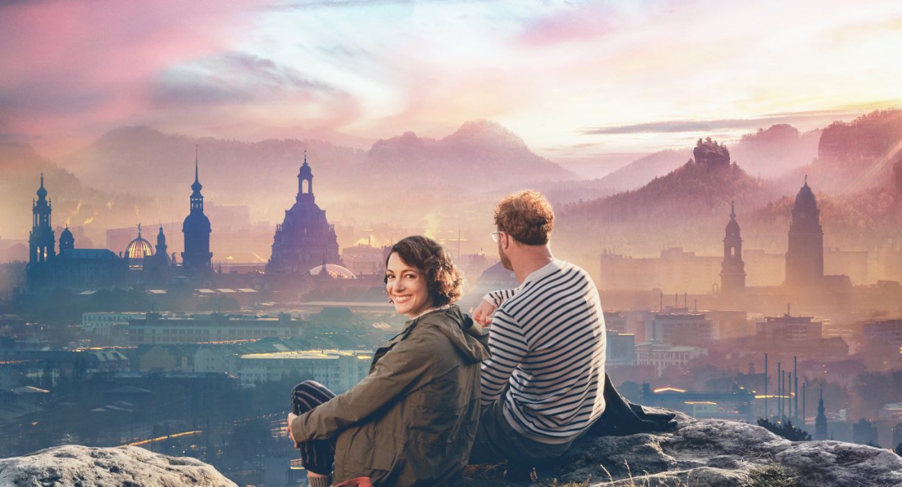 Paar auf felsigem Hügel mit Blick auf dunkle Silhouetten von Schlössern und Kirchtürmen in Dresden, der Himmel in warmes Spektrum an Farben des Sonnenuntergangs getaucht