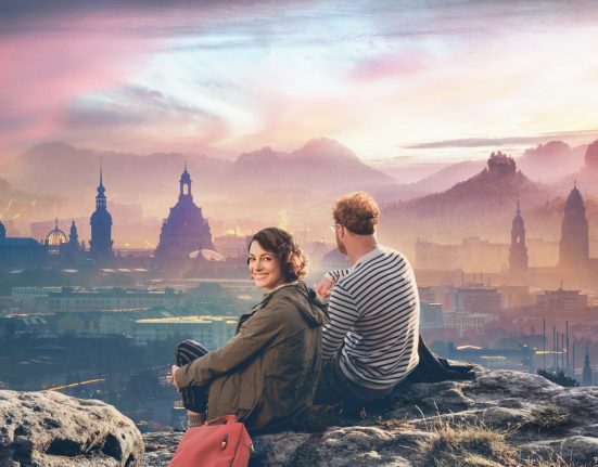Paar auf felsigem Hügel mit Blick auf dunkle Silhouetten von Schlössern und Kirchtürmen in Dresden, der Himmel in warmes Spektrum an Farben des Sonnenuntergangs getaucht