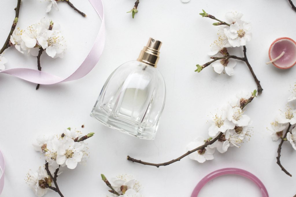 Parfumflasche liegt auf weißem Hintergrund. Aufnahme von oben, neben der Flasche Zweige mit Blüten, rosa Bänder und eine rosa Kerze