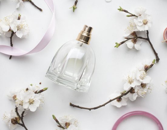 Parfumflasche liegt auf weißem Hintergrund. Aufnahme von oben, neben der Flasche Zweige mit Blüten, rosa Bänder und eine rosa Kerze