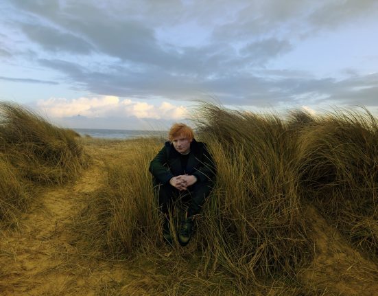 Ein Mann mit roten Haaren sitzt bei einem Strand.