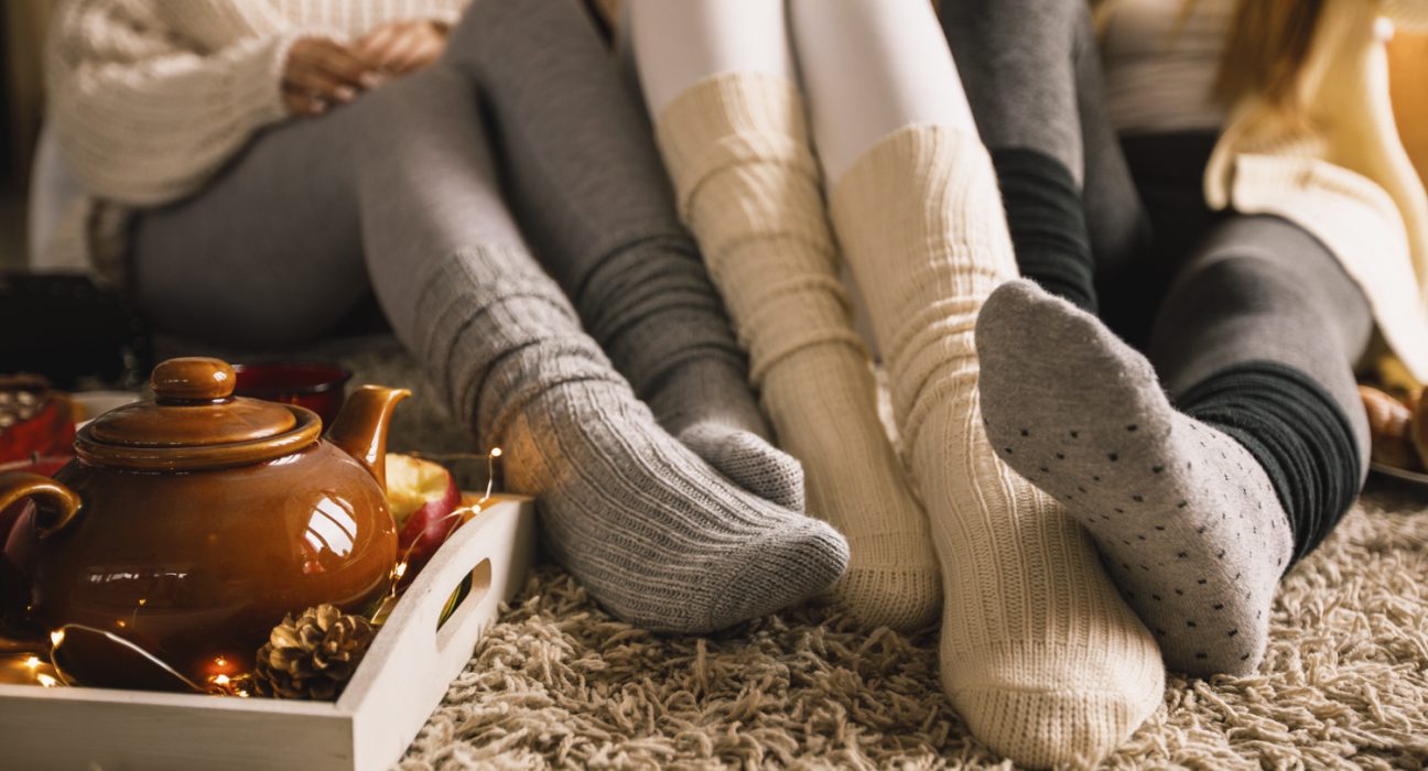 Drei Fußpaare mit kuscheligen Socken auf einem Teppich mit einer Teekanne daneben.