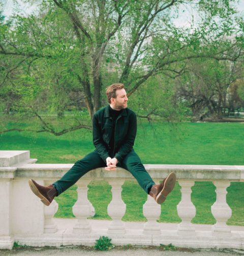Josh. in grüner Kleidung auf weißer Mauer vor einem grünem Hintergrund aus Blattwerk