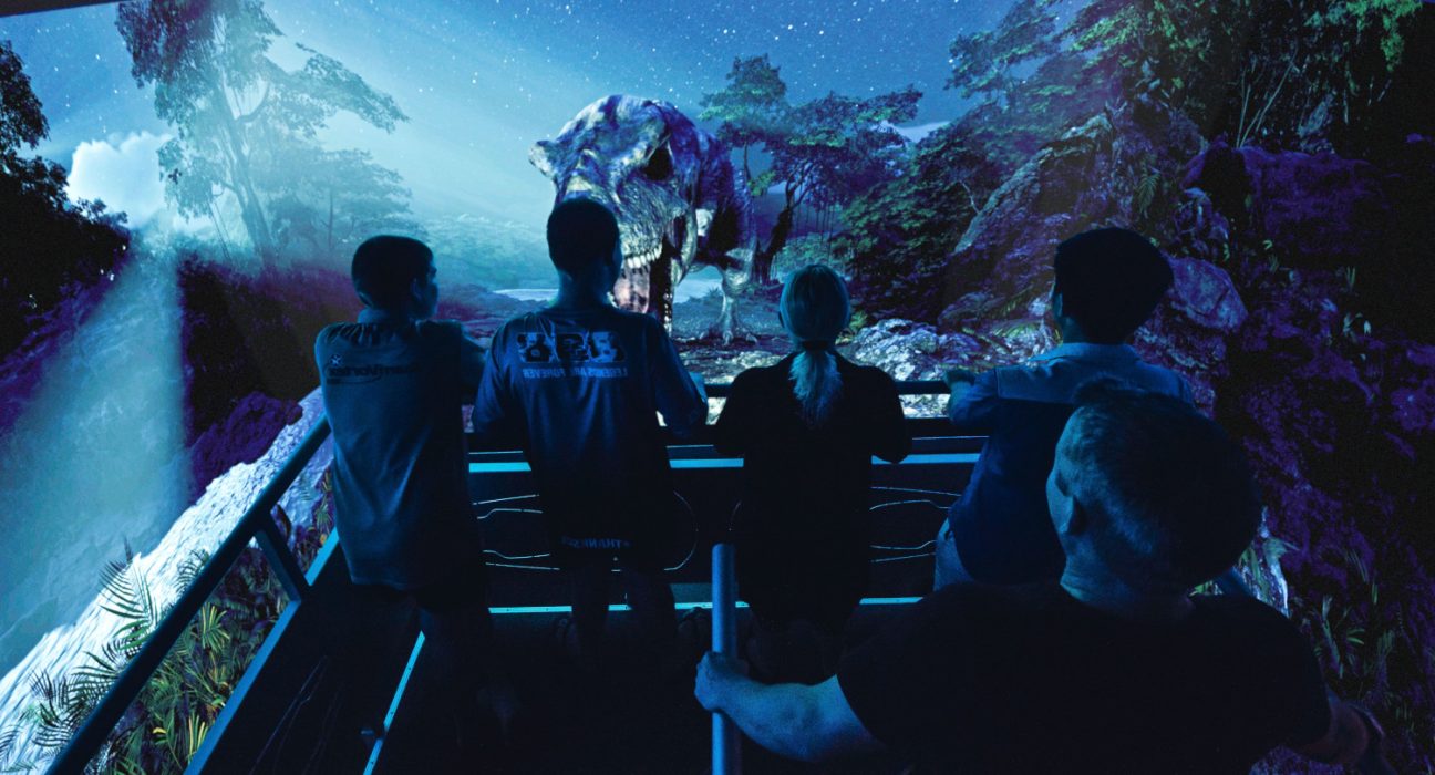 Eine Gruppe von Personen steht in einem dunklen 5D Kino und sie sehen einen Dinosaurier auf dem Bildschirm.