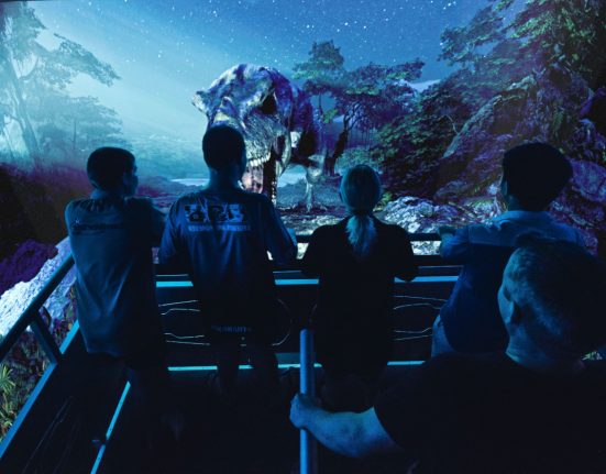 Eine Gruppe von Personen steht in einem dunklen 5D Kino und sie sehen einen Dinosaurier auf dem Bildschirm.