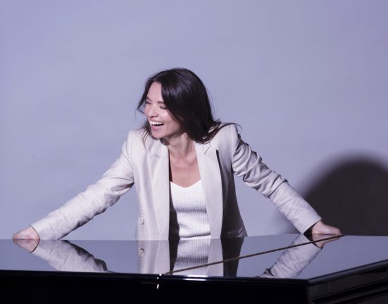 Kateryna Titova in weißem Shirt und grauem Blazer lacht mit beiden Händen auf ein Klavier gestützt