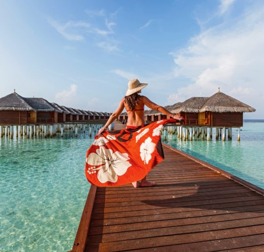 Frau in rotem Kleid und mit Hut auf einem Steg auf den Malediven