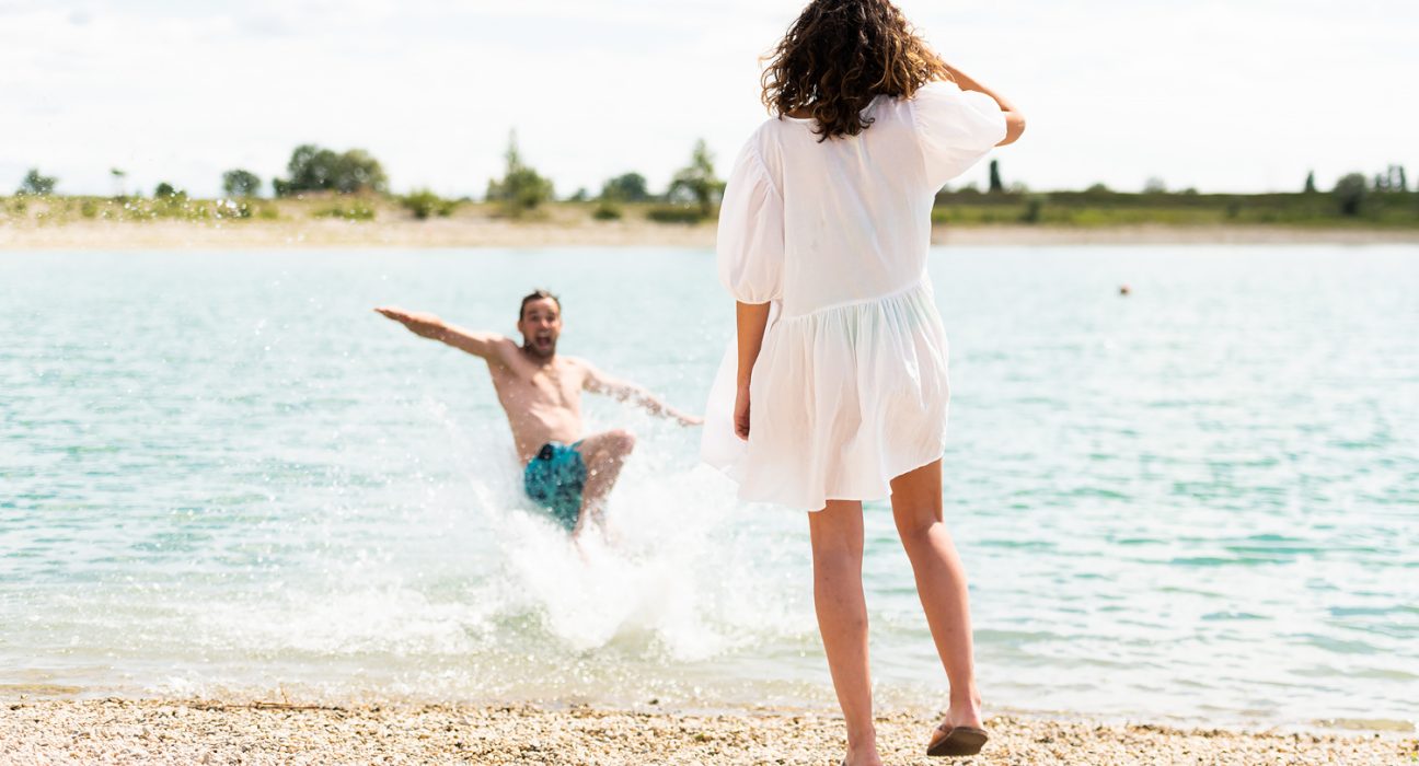 Dame in weißem Kleid beobachtet Mann in Badehose im See, sie steht mit dem Rücken zur Kamera