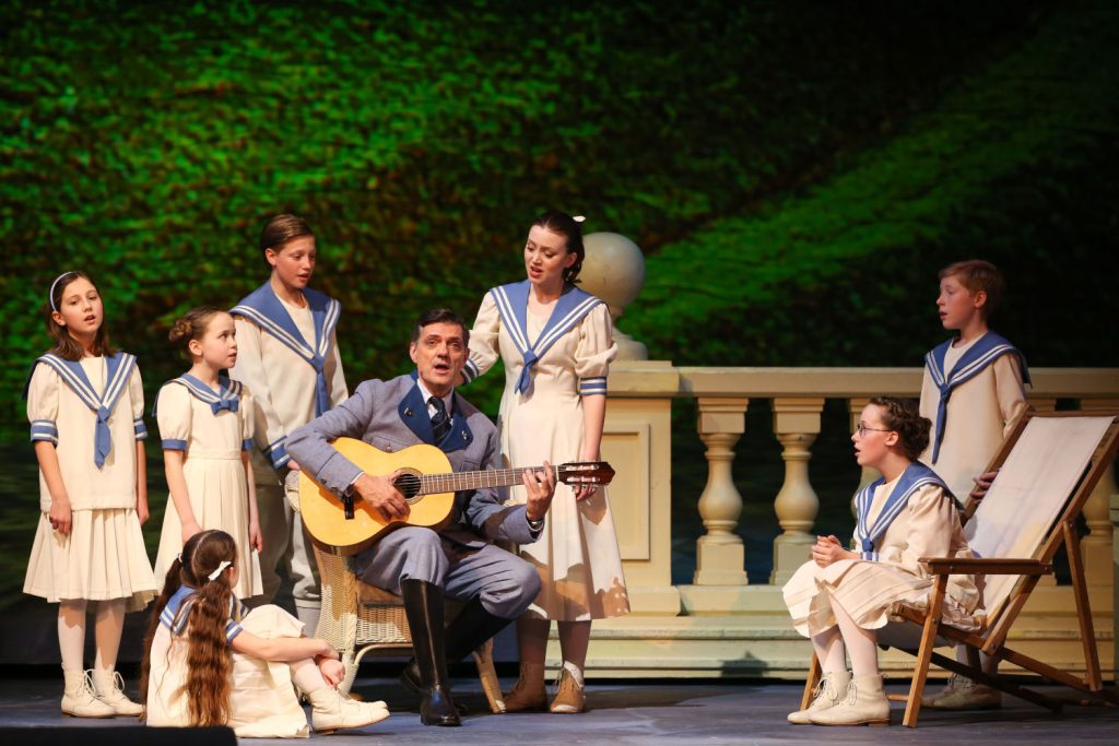 Mann mit Gitarre und Kinderchor in blau-weißen Outfits singen auf der Bühne
