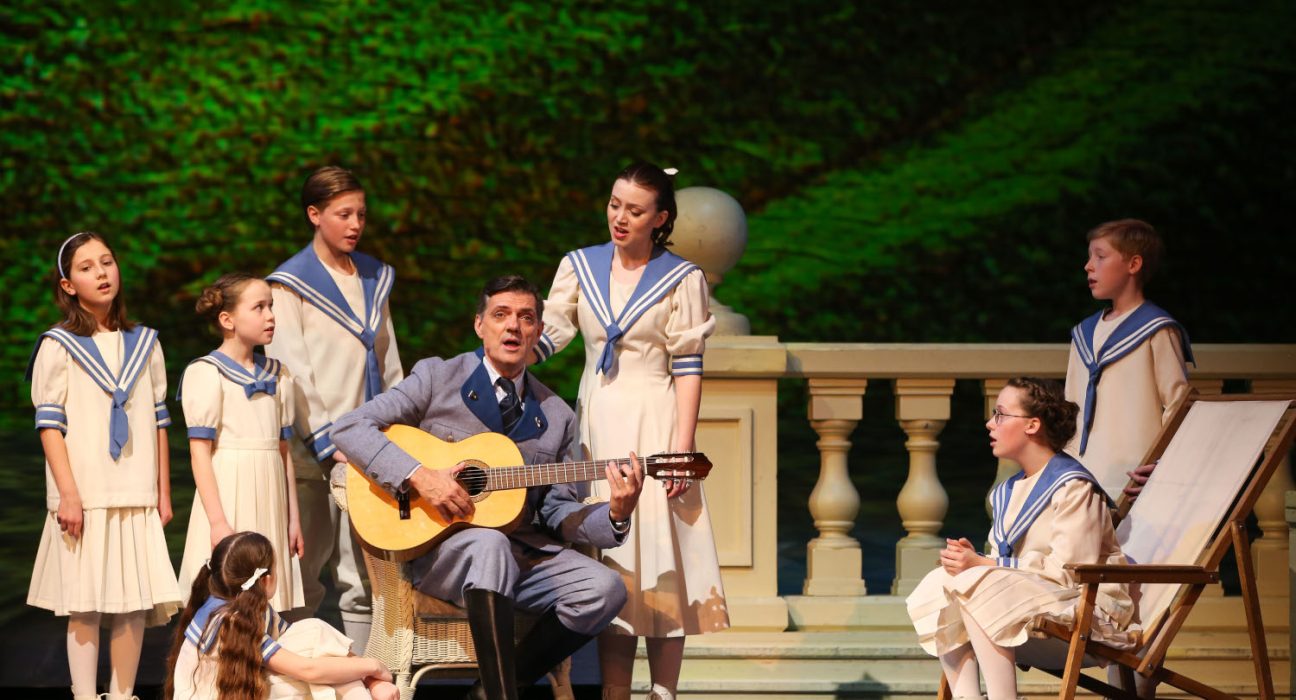 Mann mit Gitarre und Kinderchor in blau-weißen Outfits singen auf der Bühne