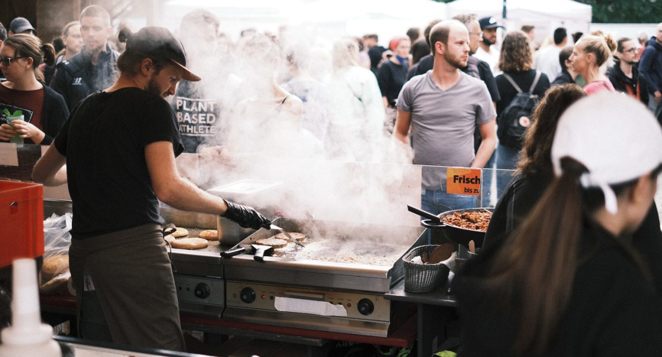 Mann grillt vegane Burger Patties beim Veganmania Streetfood Festival, Rauch steigt auf, im Hintergrund Besucher