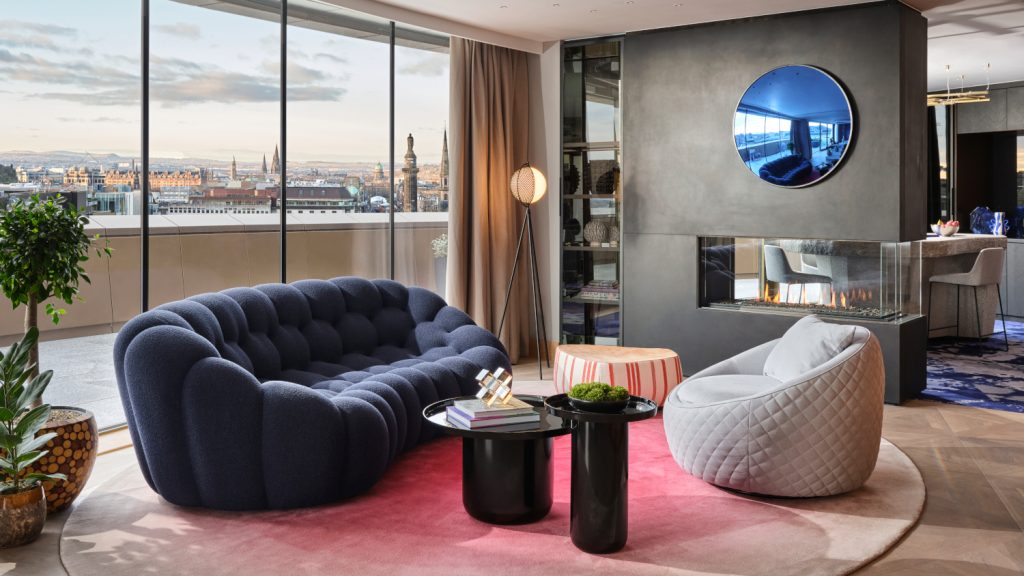 Modern eingerichtetes Wohnzimmer einer Suite des Cityhotels W Edinburgh mit roten, blauen und grauen Akzenten und Skyline von Edinburgh im Hitergrund