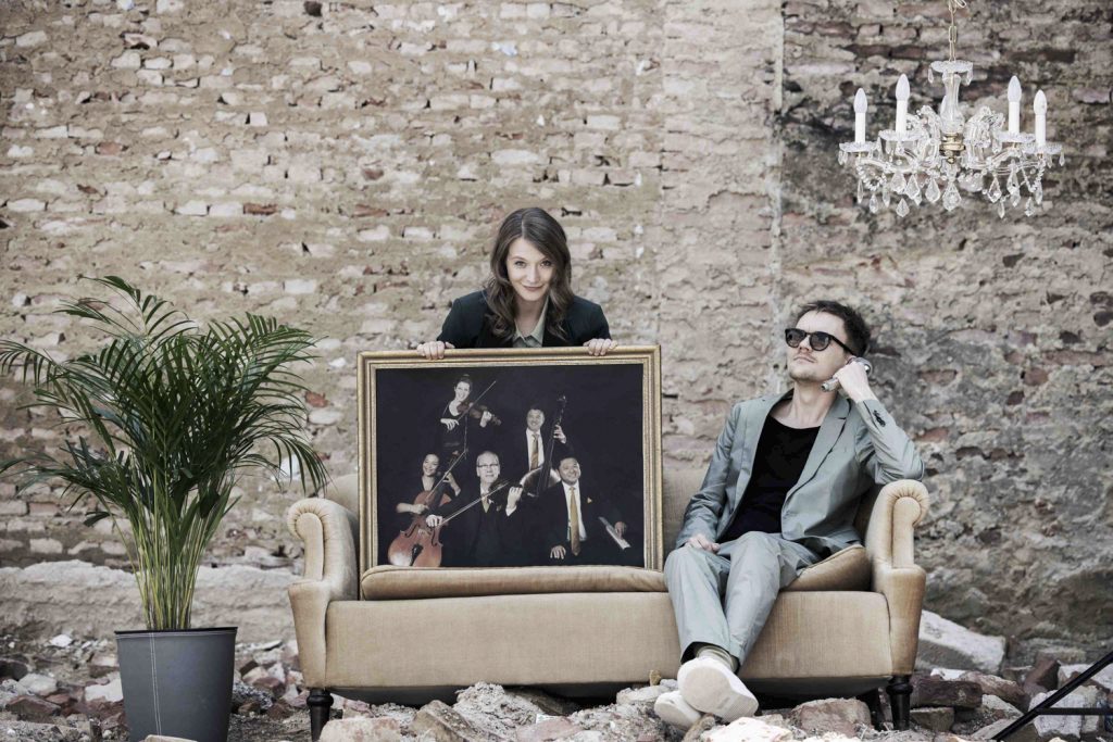 Verena Doublier und Sebastian Radon auf einer Couch vor einer grauen unverputzten Ziegelwand.