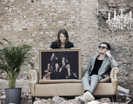 Verena Doublier und Sebastian Radon auf einer Couch vor einer grauen unverputzten Ziegelwand.