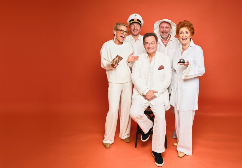 Darsteller von „Pesnion Schöller“ in ihren weißen Kostümen vor orangem Hintergrund